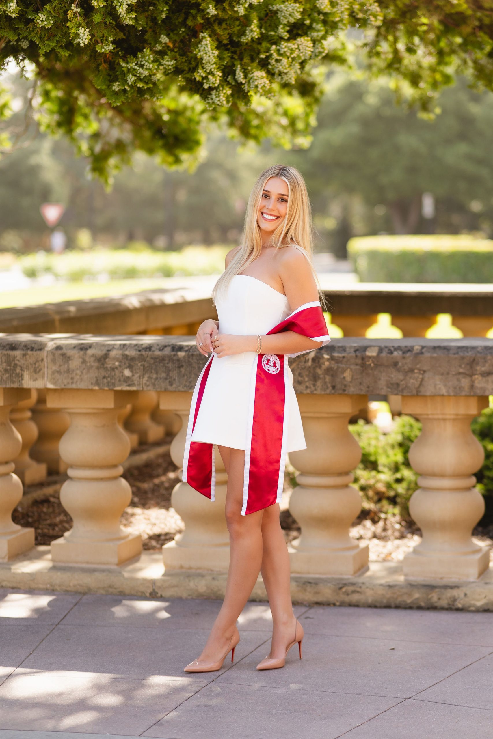 A woman in a white dress and red graduation stole stands outside by a stone railing, smiling at the camera. Trees and sunlight are in the background.